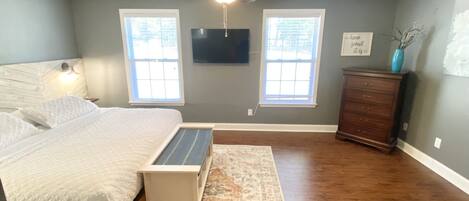 A bedroom with a bed, a desk, a dresser, a ceiling fan, and two windows.