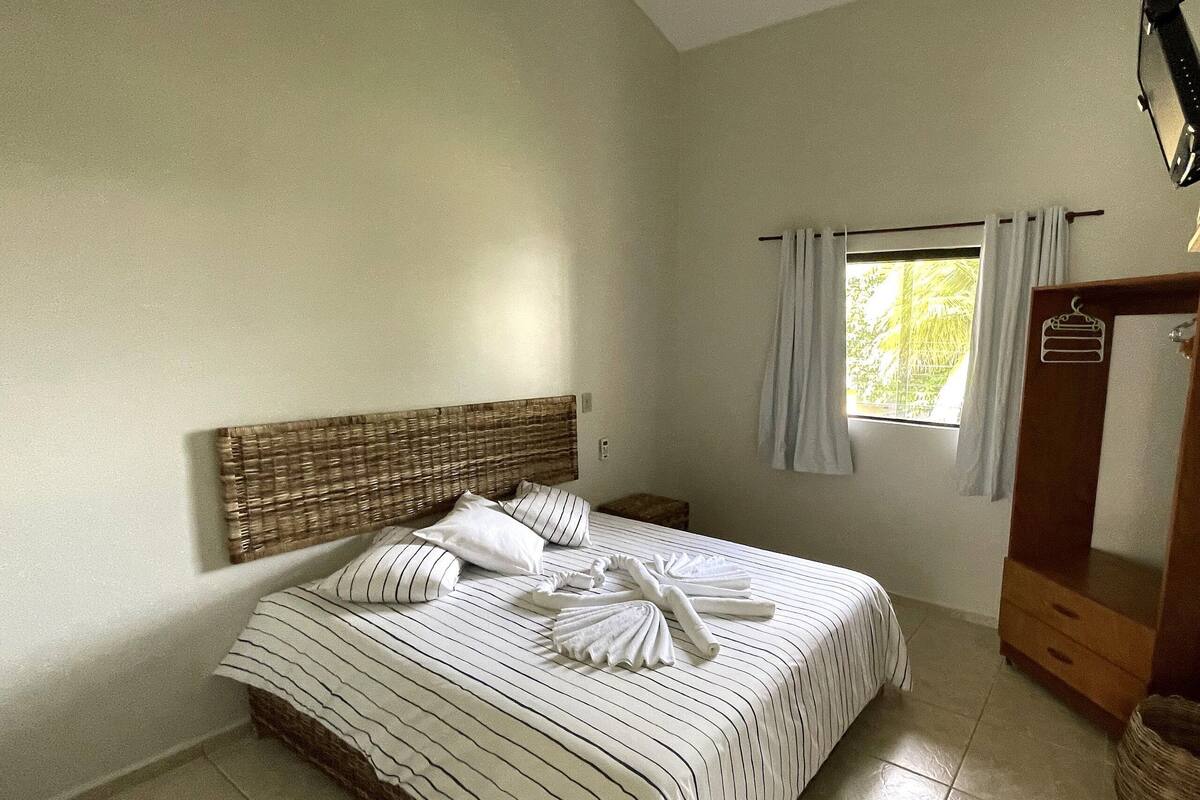 A neatly made bed with striped bedding and pillows, a wooden headboard, a small bedside table, and a window with curtains.