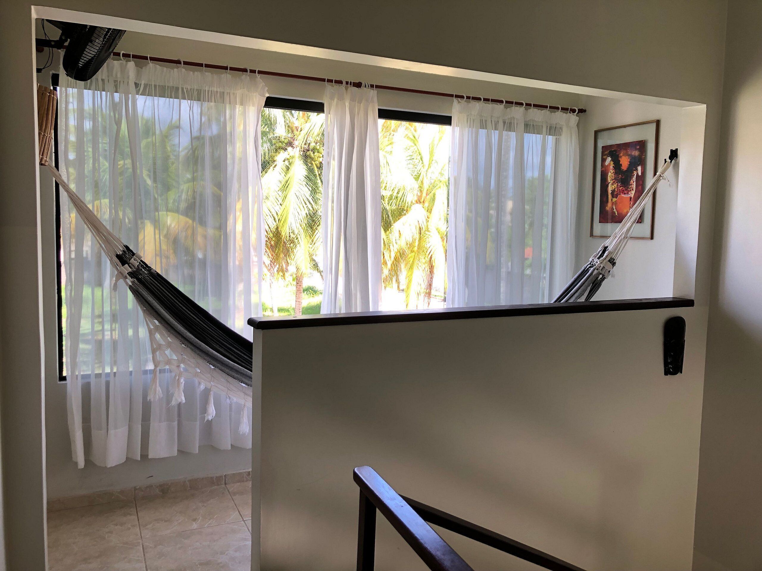 A room with a hammock, a window with sheer curtains, and a painting on the wall.