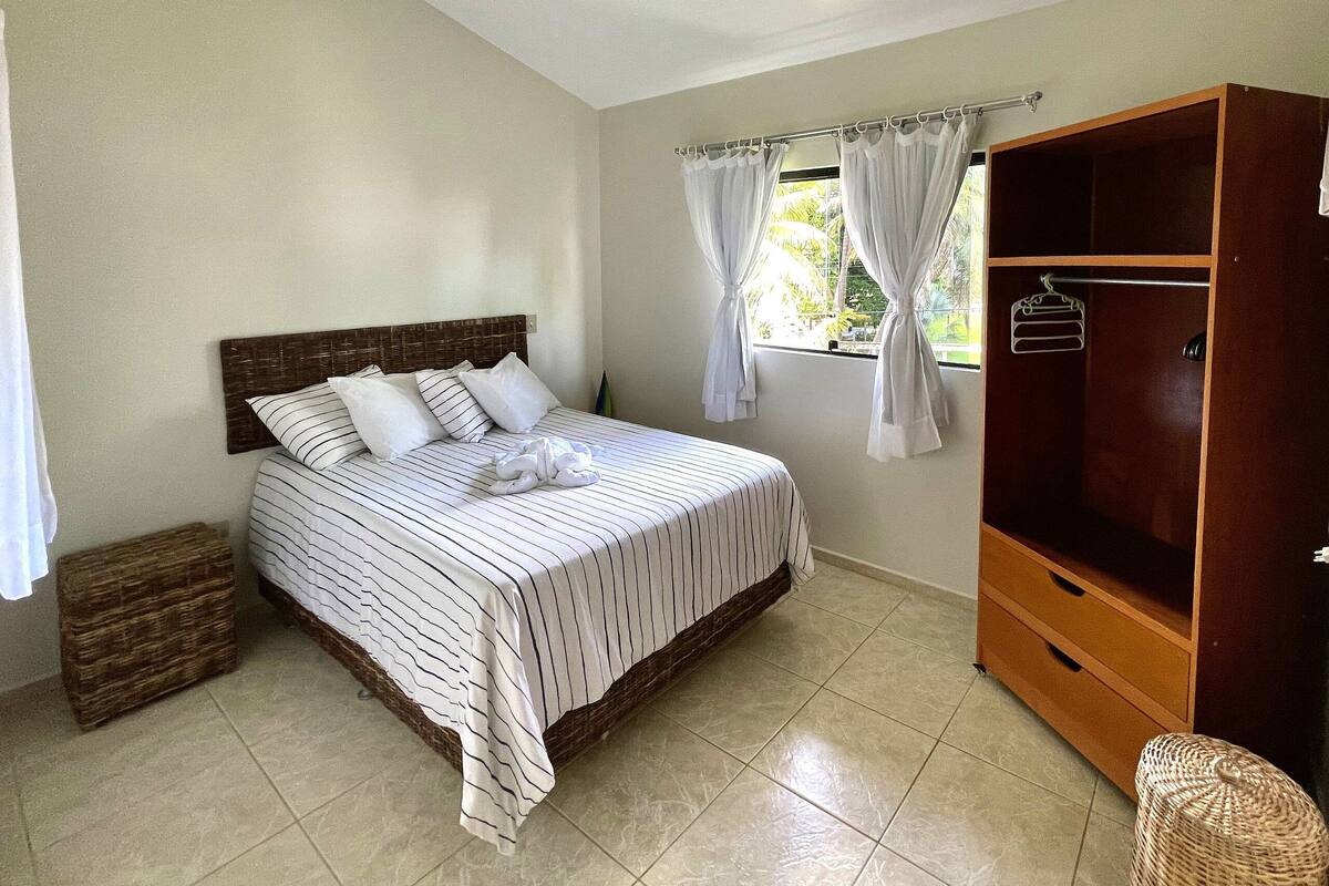 A bedroom with a bed, a wooden wardrobe, a window with curtains, and a wicker basket.