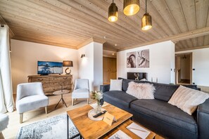 A modern living room with a dark grey sofa, two light grey armchairs, a wooden coffee table, and a wooden ceiling.