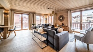 Ein geräumiger Wohnbereich mit Holzbalkendecke, großen Fenstern, einer Couch, einem Couchtisch und einem Esstisch mit Stühlen.