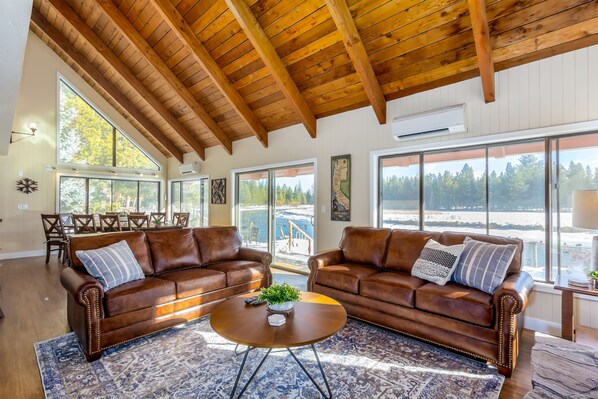 A spacious living room with two brown leather sofas, a round coffee table, and large windows offering a view of a dock and trees. - ECLI20 - River Front Home on the Deschutes, Pool Table (Sunriver)