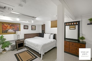 Quarto de hotel com cama, mesa de cabeceira, luminária, cômoda, espelho e um quadro na parede.