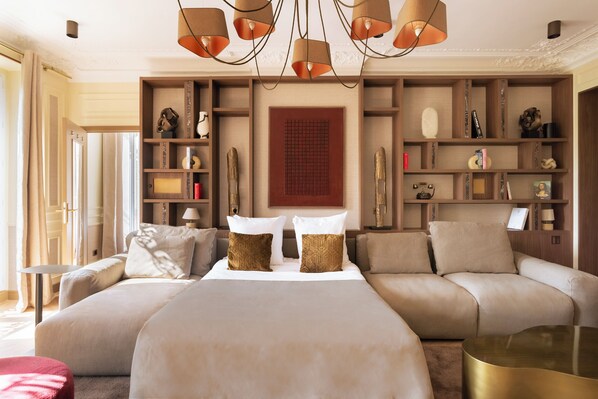 A spacious bedroom with a large bed, a sofa, and a small table. There are shelves with decorative items and a painting on the wall.
