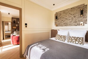 A bedroom with a bed, a red chair, a wooden headboard, a wall-mounted light fixture, and a textured wall. - Merveil - Luxury Suite - Marais - Ponceau (Paris)