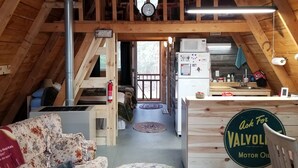 Una habitación acogedora en el ático con vigas de madera, un ventilador de techo, un reloj, una cama, un sofá, una zona de cocina y un cartel de Valvolo Motor Oil.