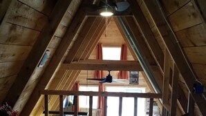 Un dormitorio en el ático con techo inclinado, vigas de madera, ventilador de techo, cama con colcha estampada y vista de la habitación contigua a través de una ventana.