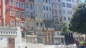 Straßenansicht mit mehrstöckigen Gebäuden, einem sichtbaren Geschäft mit der Beschriftung 'Catarino' und Außenbestuhlung mit Sonnenschirmen.