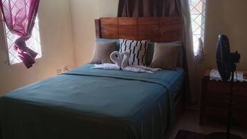 A bedroom with a bed, a nightstand, a fan, and a window with curtains.