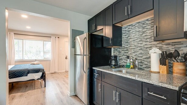 A compact kitchen with dark cabinets, a stainless steel refrigerator, and a tiled backsplash. - Melrose Studio - Pool & Free Parking Near Downtown (Nashville)