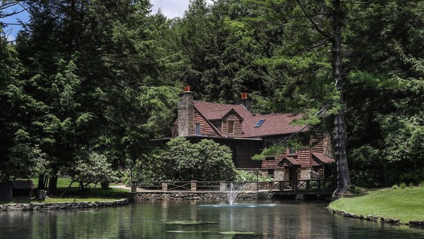 A rustic lodge with a stone chimney, a wooden deck, and a pond with a fountain. - Fin & Feather at Seven Springs (Champion)