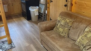 Un acogedor interior de cabaña con suelo de madera, un sofá con cojines estampados, una zona de cocina con estufa y cafetera, y una ventana con cortinas.