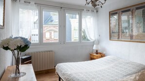Une chambre à coucher avec un grand lit, une table de chevet, une lampe, un vase de fleurs et une fenêtre avec des rideaux.