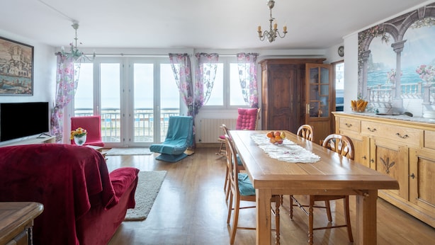 Un espace repas avec une table en bois, des chaises et une vue sur la mer grâce à de grandes fenêtres.