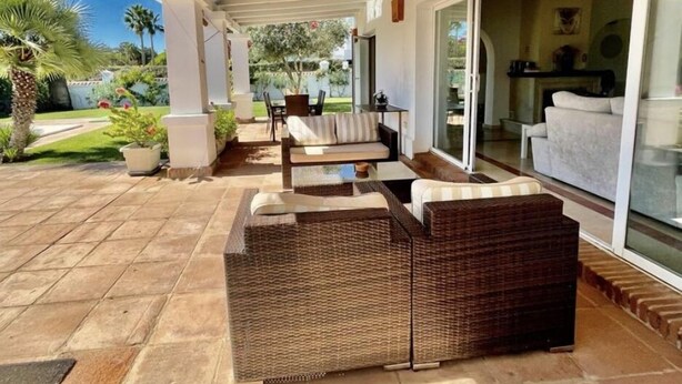 A covered patio area with wicker furniture, a glass door leading to a living room, and a garden with palm trees. - 5star A/C villa, 5 min beach & marina w bars refurb. pool & garden.  (Cadiz)