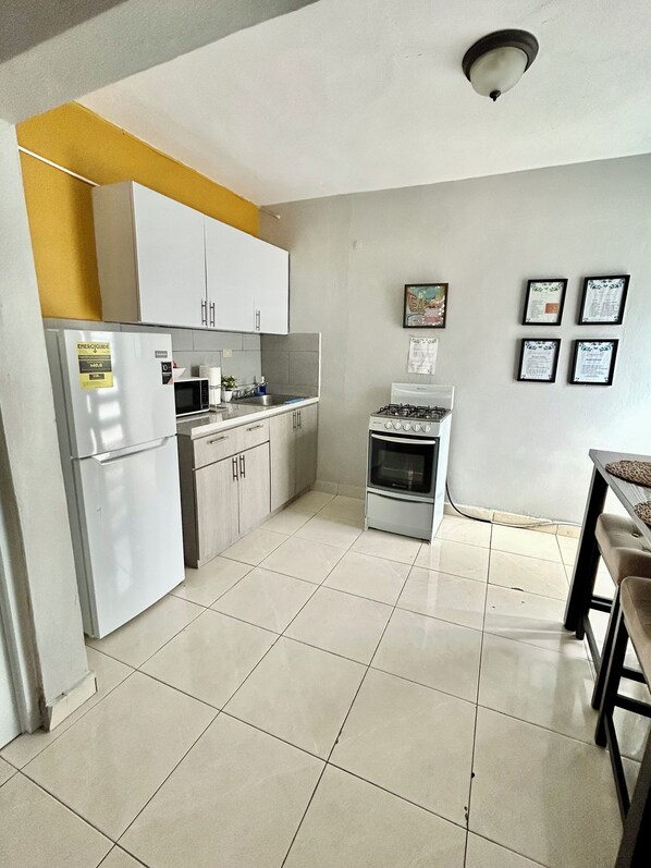 Una cocina compacta con armarios blancos, un refrigerador, un microondas y una estufa. Se observan certificados enmarcados en la pared y un pequeño comedor con una mesa y sillas.