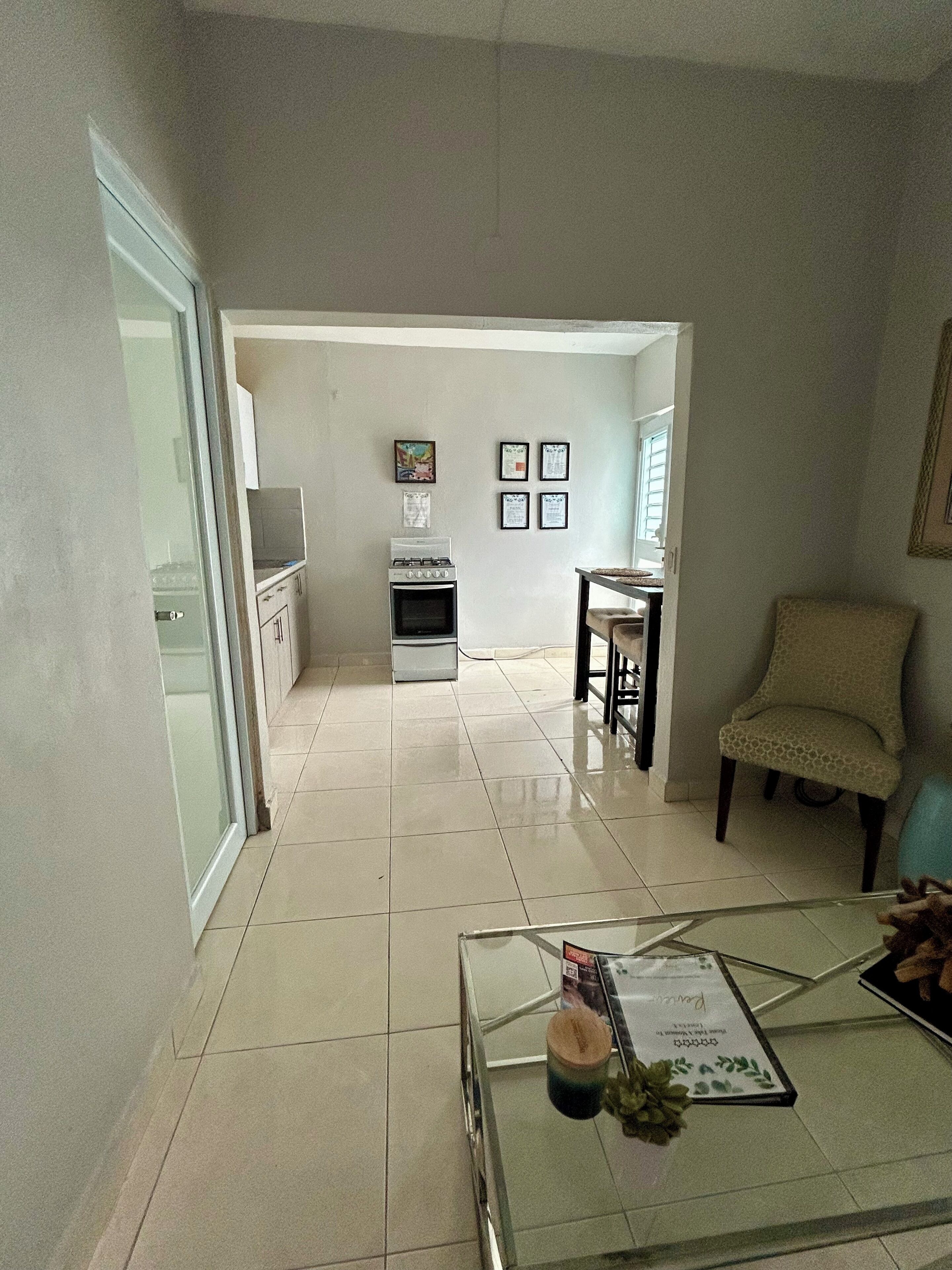 A spacious living room with a glass coffee table, a sofa, and a dining area with a chair and table. A kitchen is visible in the background with a stove and cabinets.