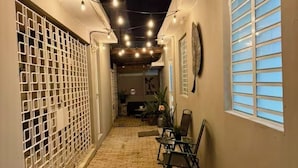 Un étroit couloir en briques dont le plafond est décoré de guirlandes lumineuses, avec une porte métallique sur la gauche et une fenêtre avec des stores sur la droite.
