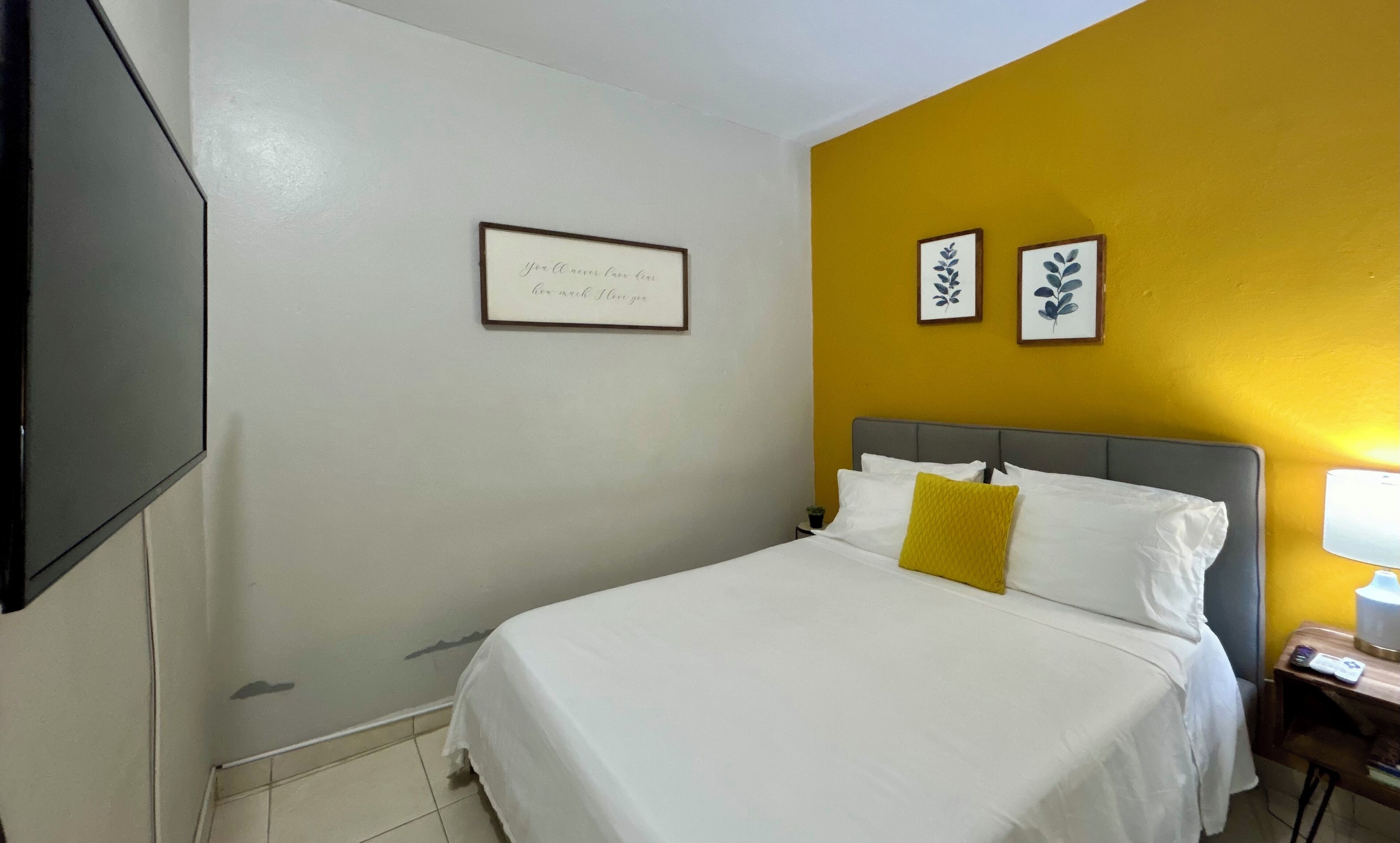 A hotel room with a bed, a bedside table, a lamp, and a framed quote on the wall.