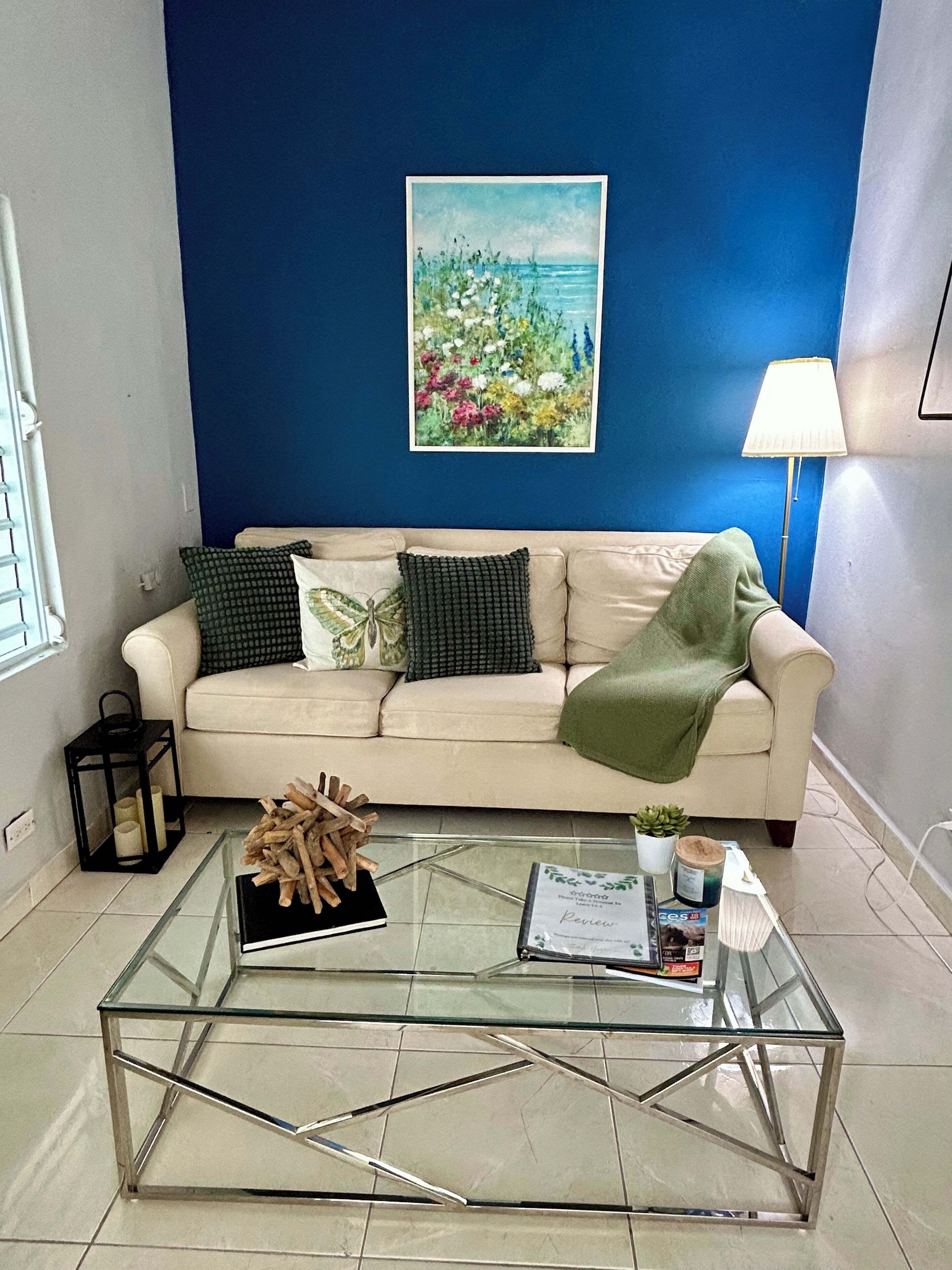 A living room with a beige sofa, a glass coffee table, a painting of flowers, and a lamp.