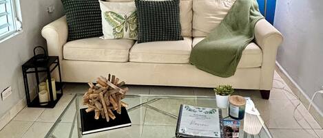 A living room with a beige sofa, a glass coffee table, a painting of flowers, and a lamp.
