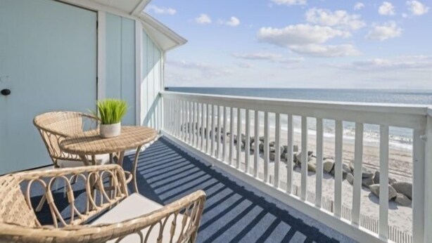 A balcony with wicker furniture and a potted plant, overlooking a beach with a white fence and rocks. - A Little Shore Thing 1 Bedroom Condo by RedAwning (Carolina Beach)