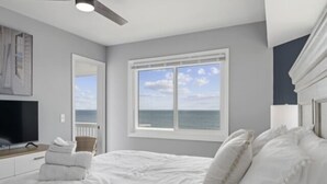 A bedroom with a bed, a television, a window with a view of the ocean, and a ceiling fan. - A Little Shore Thing 1 Bedroom Condo by RedAwning (Carolina Beach)