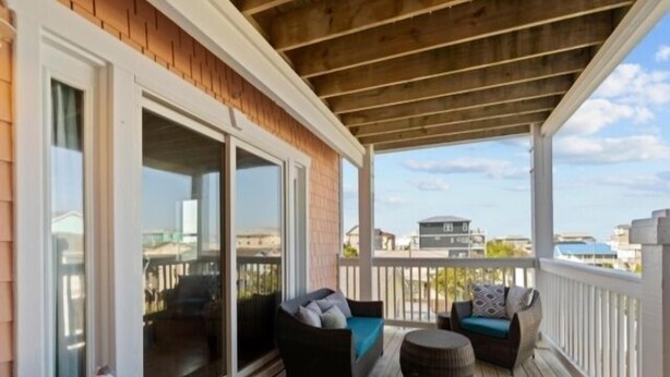 A covered outdoor deck with wooden flooring, two sofas with cushions, and a round ottoman. - Almost Heaven 4 Bedroom Home by RedAwning (Carolina Beach)
