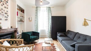 Un salon avec un canapé gris foncé, un fauteuil vert, une petite table blanche et une chaise en osier. On y trouve une cheminée, une bibliothèque et une lampe fixée au mur.