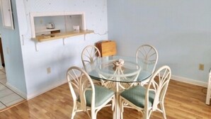 Ein Essbereich mit einem runden Glastisch, um den weiße Korbstühle mit grünen Kissen stehen. An der Wand über dem Tisch ist ein Holzregal mit Küchenutensilien angebracht.