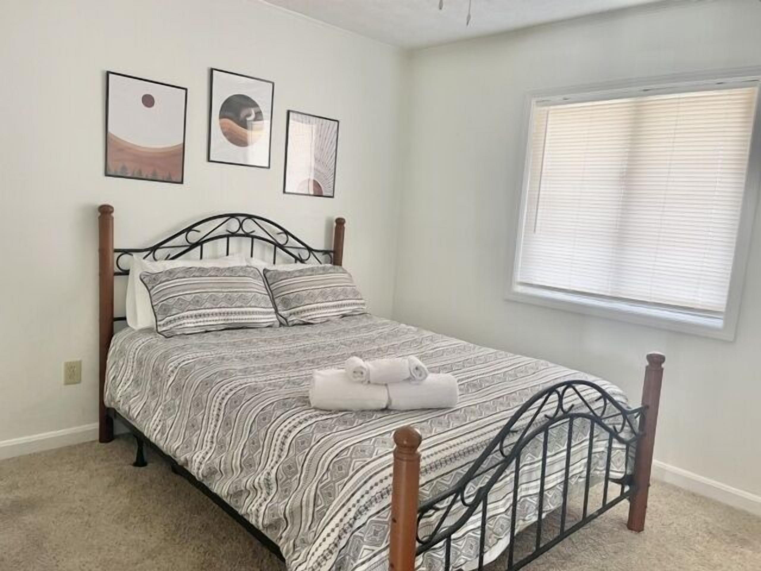 Una cama bien tendida con una colcha estampada y almohadas, una ventana con persianas, y tres cuadros en la pared.