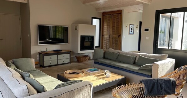 A modern living room with a sofa, a coffee table, a flat-screen TV, a fireplace, and a large window. - Villa Les Sables, Ile de Ré, a few minutes from the ocean (Sainte-Marie-de-Ré)