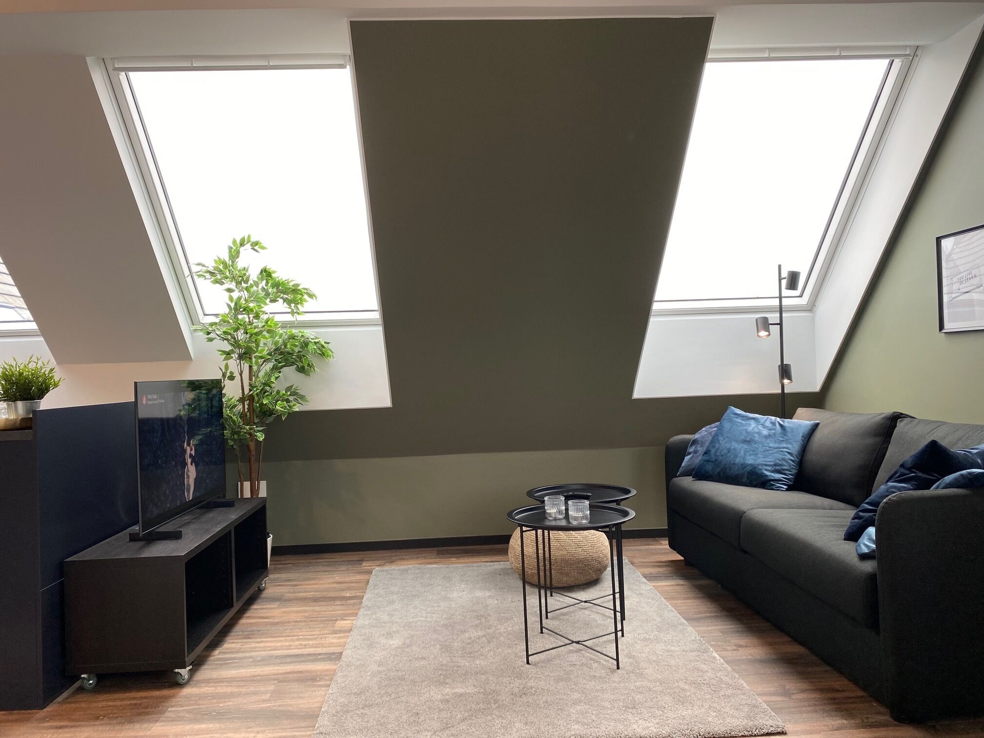 A modern living room with a dark sofa, a small round table, and a TV on a stand. There are two large windows providing natural light.