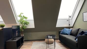 A modern living room with a dark sofa, a small round table, and a TV on a stand. There are two large windows providing natural light.