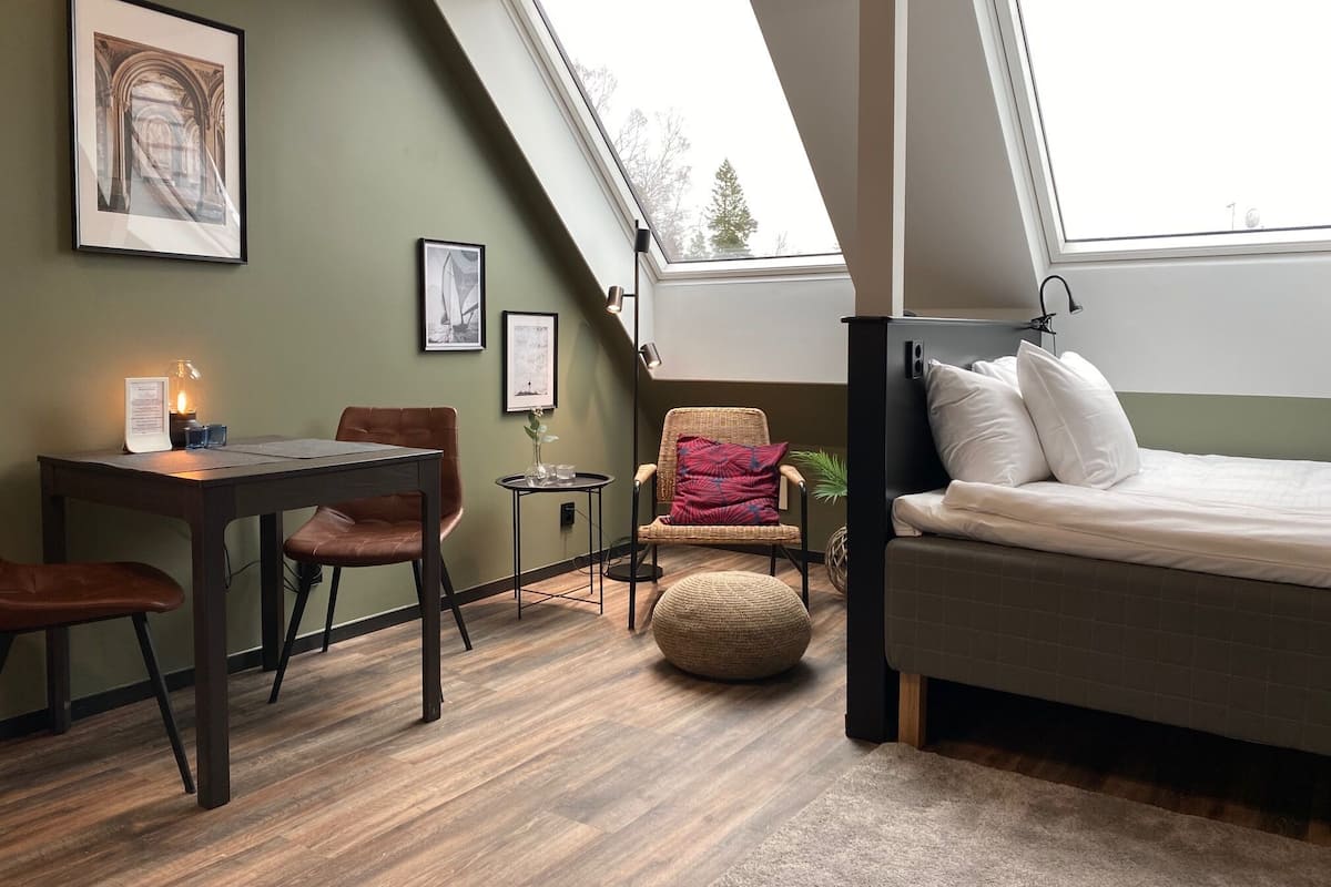 A cozy room with a bed, a desk, and a chair. There's a skylight above the bed and a framed picture on the wall.