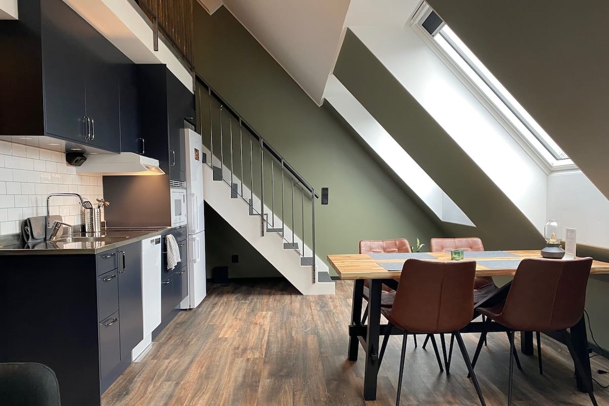 A modern kitchen with dark cabinetry, a wooden dining table, and a staircase leading to a loft area.
