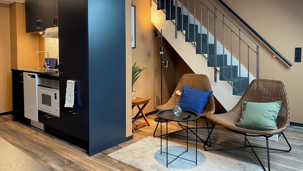 A modern living room with a kitchen area, a staircase, and two chairs with cushions.