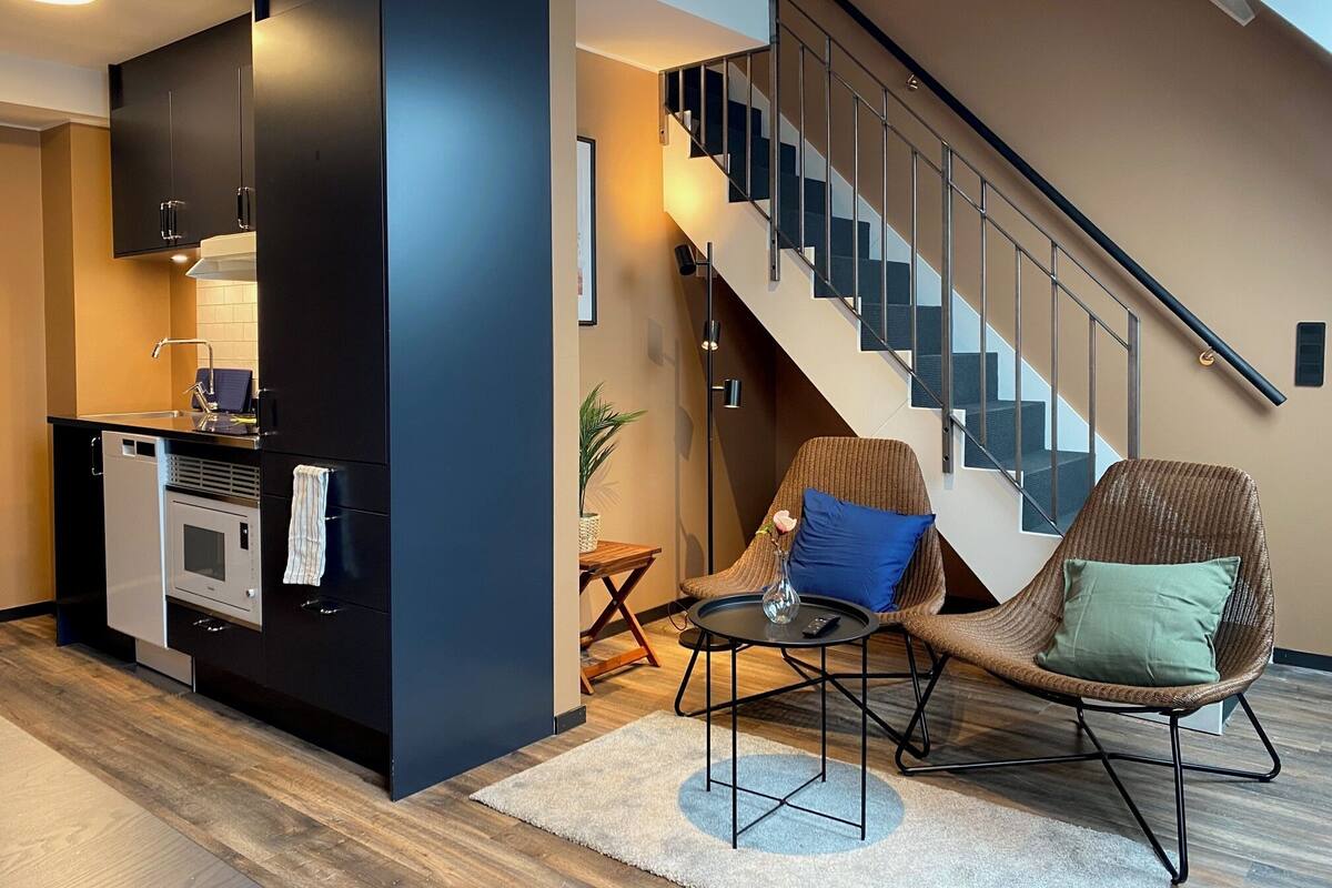 A modern living room with a kitchen area, a staircase, and two chairs with cushions.