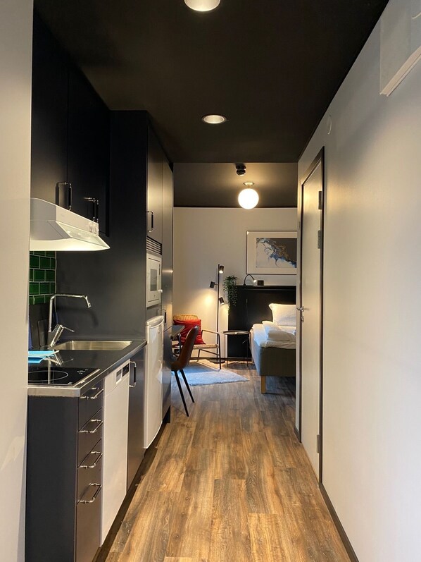 A narrow hallway with a kitchenette on the left, a dining area with a chair and table, and a bed with a nightstand in the background. - Magnolia House Sollentuna (Sollentuna)