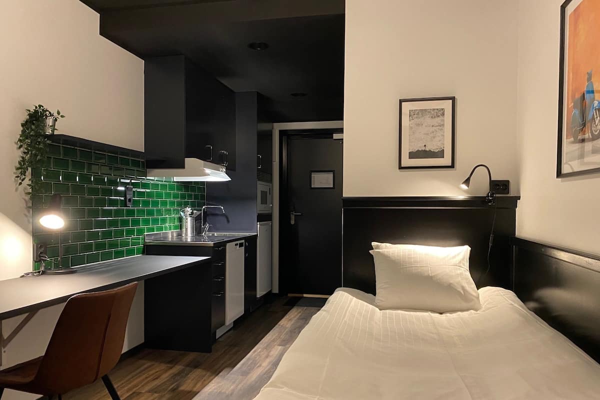 A compact hotel room with a kitchenette, a bed with white bedding, a desk with a chair, and a small potted plant.