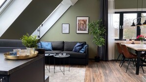 A modern living space with a loft-style ceiling, a dark-colored sofa with blue cushions, a small round table, and a dining area with a wooden table and chairs.