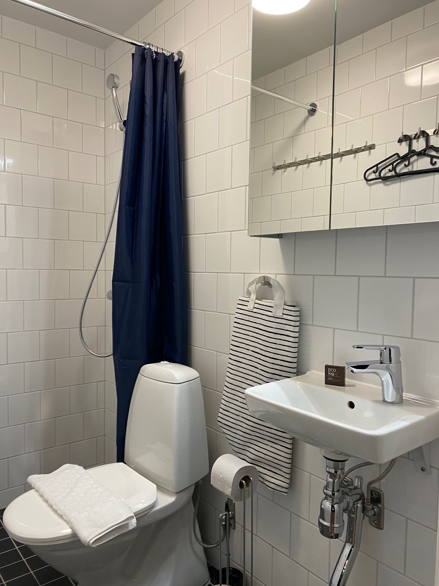 Un baño con inodoro, lavamanos, ducha con cortina azul y espejo con toallero.