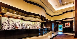 A modern hotel lobby with a curved reception desk, a large abstract wall art piece, and a digital display screen.