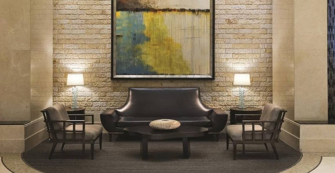 A lobby with a stone wall, a large abstract painting, a leather sofa, two side tables with lamps, and four chairs.