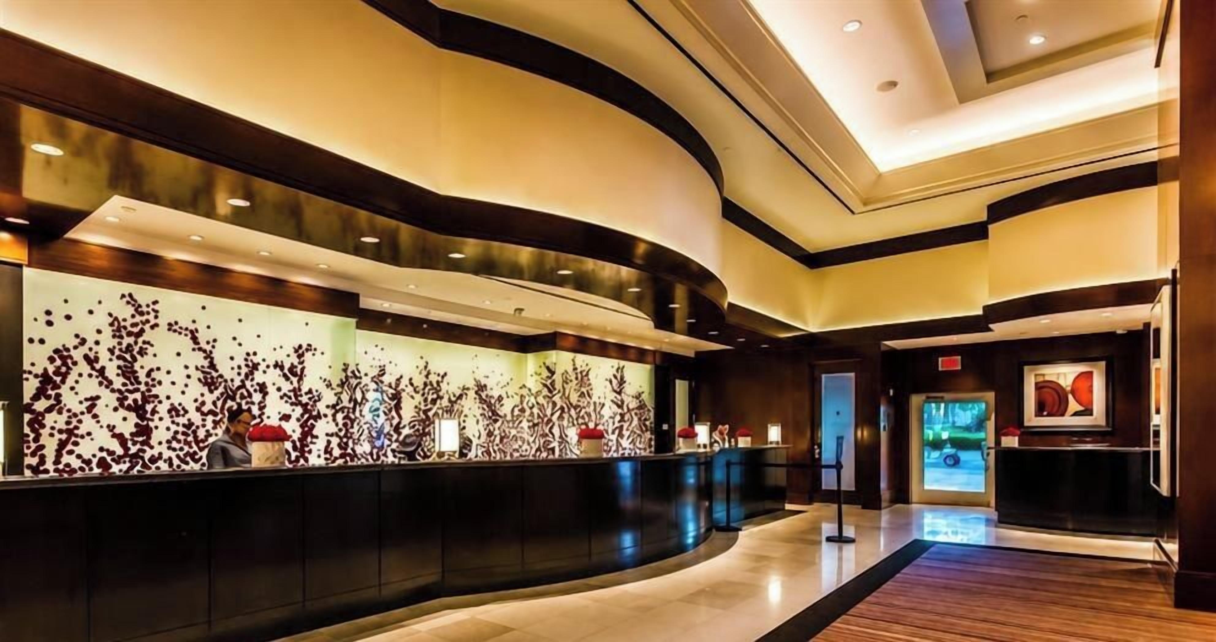 Een moderne hotellobby met een gebogen receptiebalie, een groot abstract kunstwerk aan de muur en een digitaal beeldscherm.