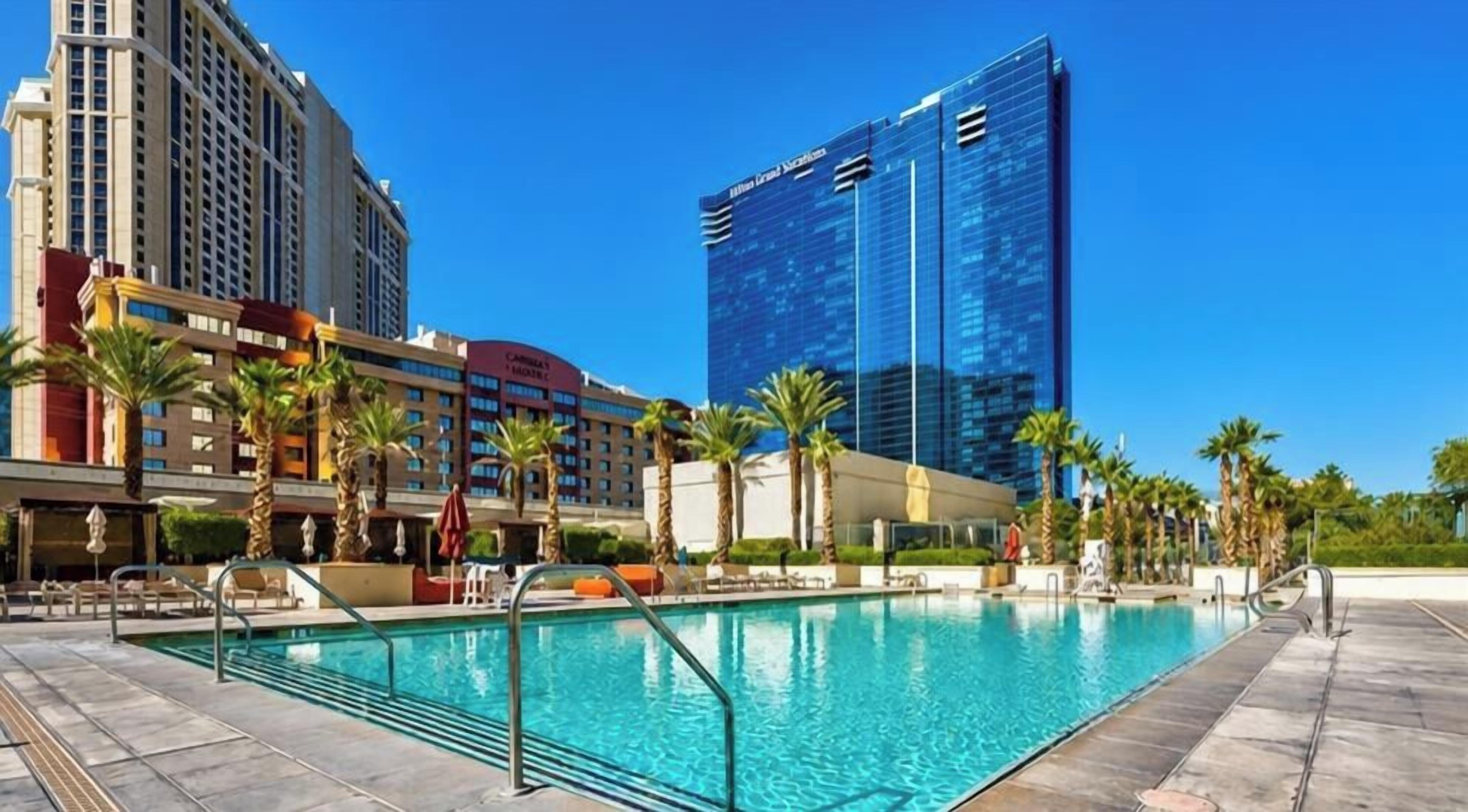 Un espace piscine d’hôtel avec une piscine à l’eau d’un bleu limpide, des palmiers et des bâtiments modernes en arrière-plan.