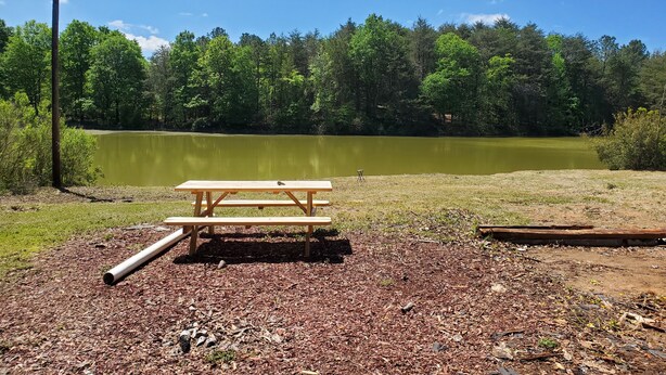 A picnic table and benches in a park with a lake and trees in the background. - Emmar Blue + Fishing Lake/ Pond  (Charlotte)