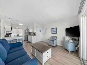A modern living room with a sofa, a TV, a small table, and a kitchen area in the background.