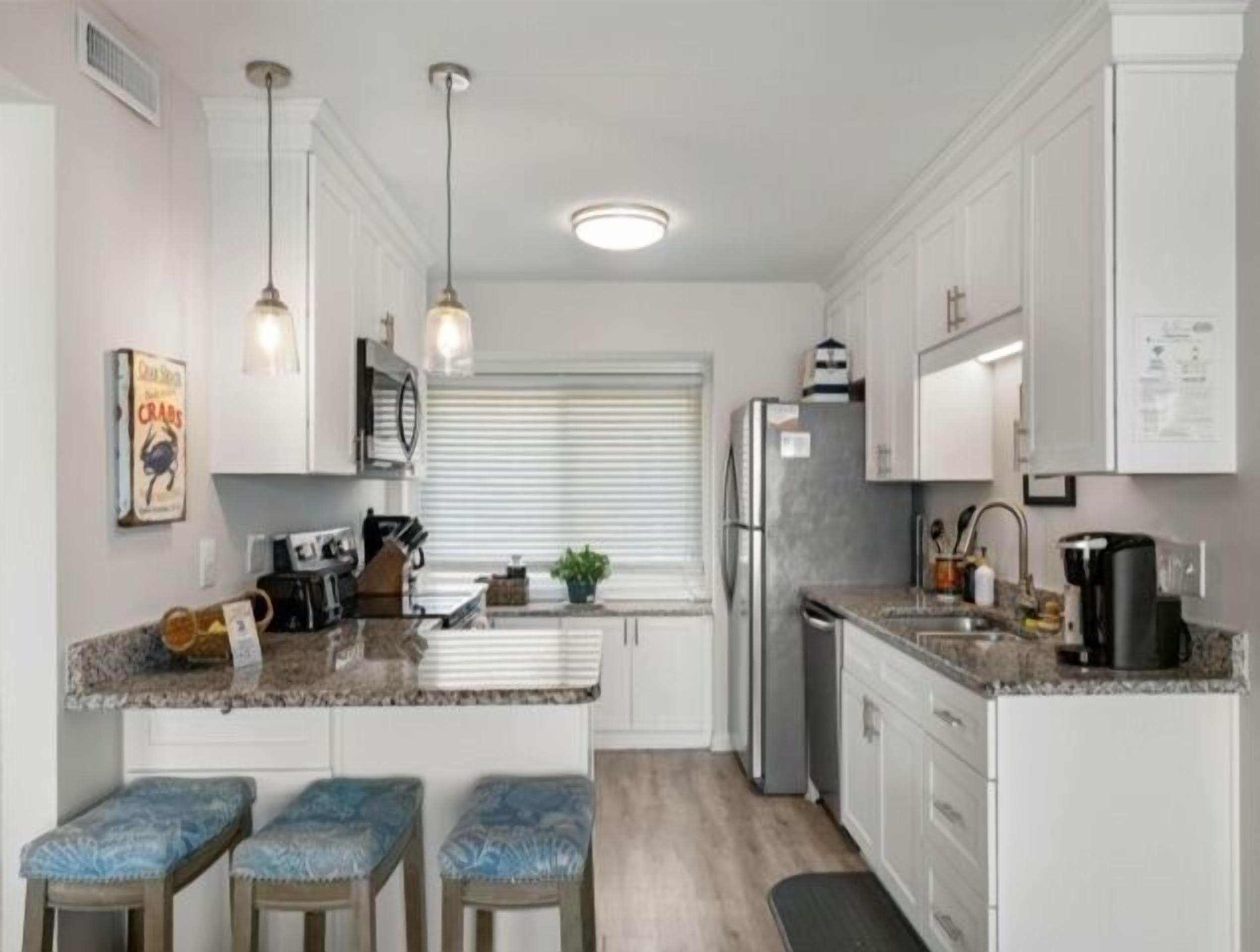 A modern kitchen with white cabinets, a granite countertop, a central island with stools, pendant lighting, and a refrigerator.
